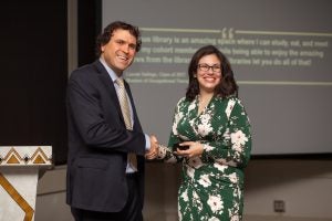 Two people shaking hands in front of a presentation screen. One is in a dark suit, the other in a green floral dress.