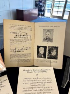 An open book on display shows a page with a heredity chart and another page with images of individuals.