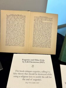 An open book titled "Eugenics and Other Evils" by G.K. Chesterton (1927) is displayed with a printed card below it summarizing the book's critique of eugenics.